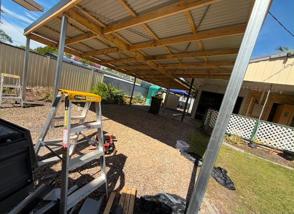 Timber-framed carport with steel posts and sheet roofing over a gravel driveway beside a brick home
